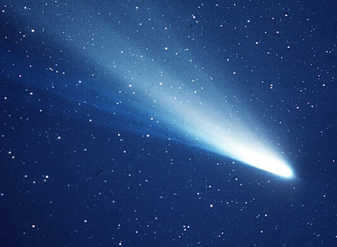 A striking image of a brilliant comet streaking across a deep blue, star-studded sky. The comet's nucleus is intensely bright, fanning out into a long, glowing tail that gradually diffuses into the darkness, with numerous small white stars scattered throughout the celestial backdrop.
