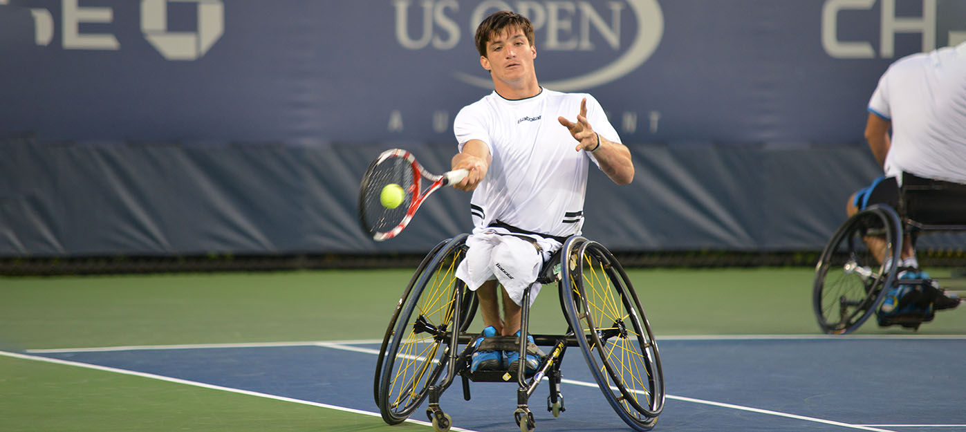 A person in a wheelchair hits a tennis ball with a racket while on a tennis court. The U.S Open logo is in the  background.